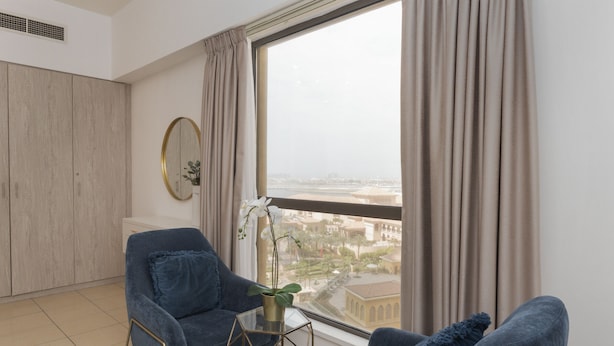A modern living room with a large window, a sofa, a small table, and a decorative plant. - Cozy JBR 2BR Retreat Steps to the Beach The Walk (Dubai)