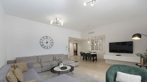 A modern living room with a sectional sofa, a round coffee table, a green armchair, a dining area with chairs, a TV, and a wall clock. - Cozy JBR 2BR Retreat Steps to the Beach The Walk (Dubai)