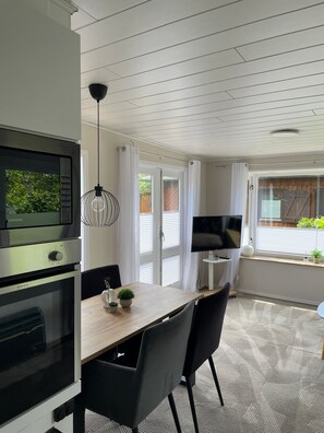 A modern kitchen with a built-in oven, a dining table with chairs, a TV, and a window with curtains.