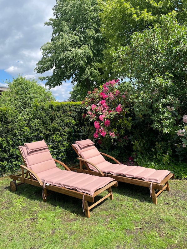 Two wooden lounge chairs with cushions, positioned on a grassy lawn with dense green foliage and flowering plants in the background.