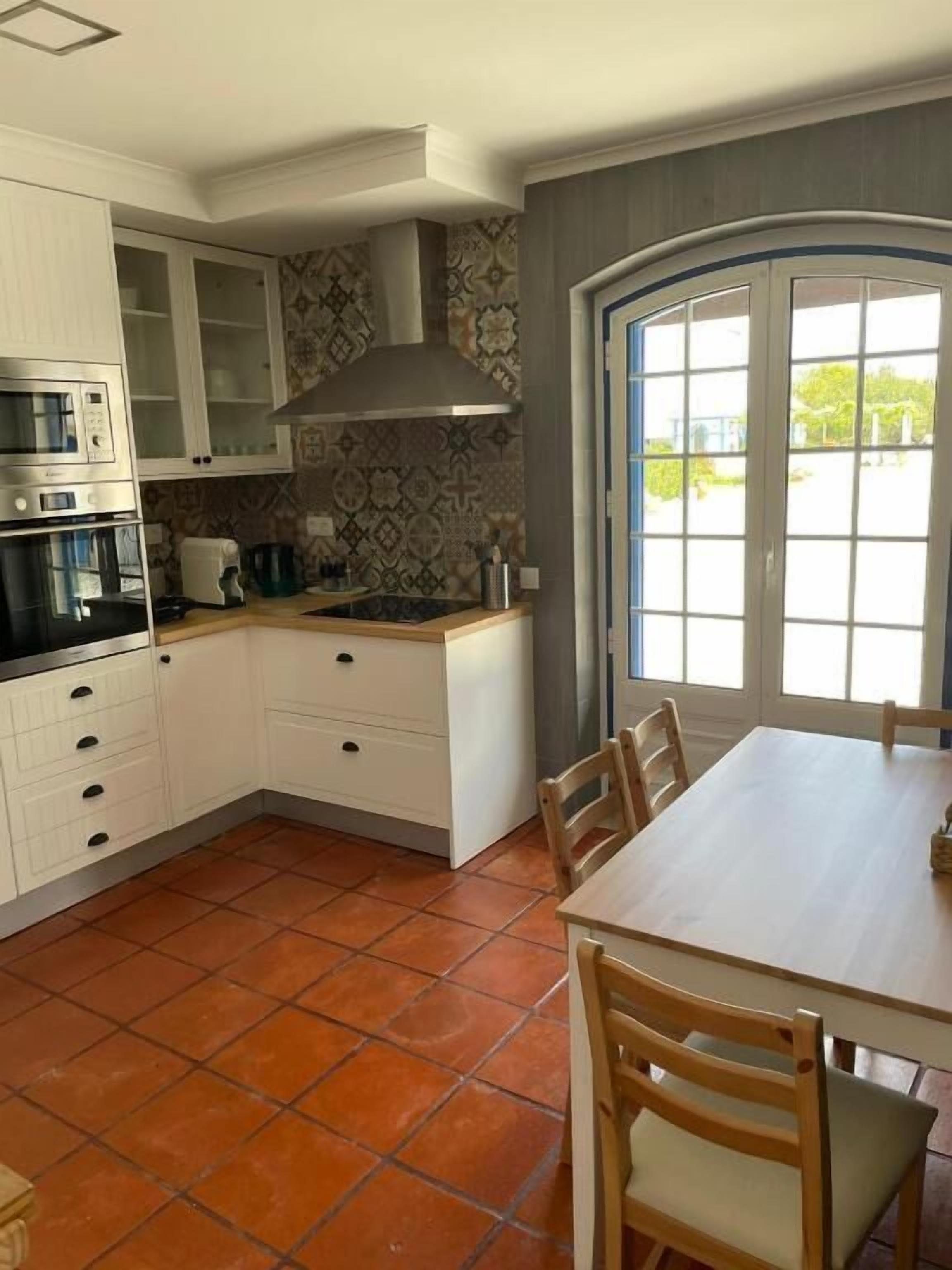 Een keuken met witte kastjes, een fornuis en een spoelbak. Er staat een eettafel met stoelen.