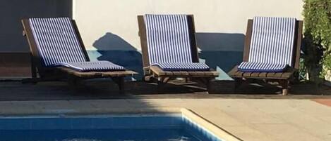 A swimming pool with three striped lounge chairs and a small window with a light fixture.