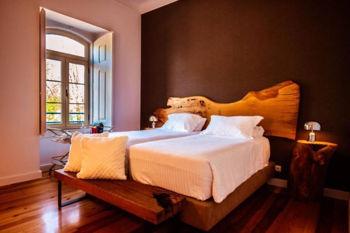 A double bed with white bedding, a wooden headboard, a bedside table with a lamp, and a window with a view of greenery.