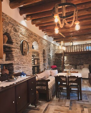 A rustic interior with exposed stone walls, wooden beams, a stone fireplace, a bed, and a dining area.