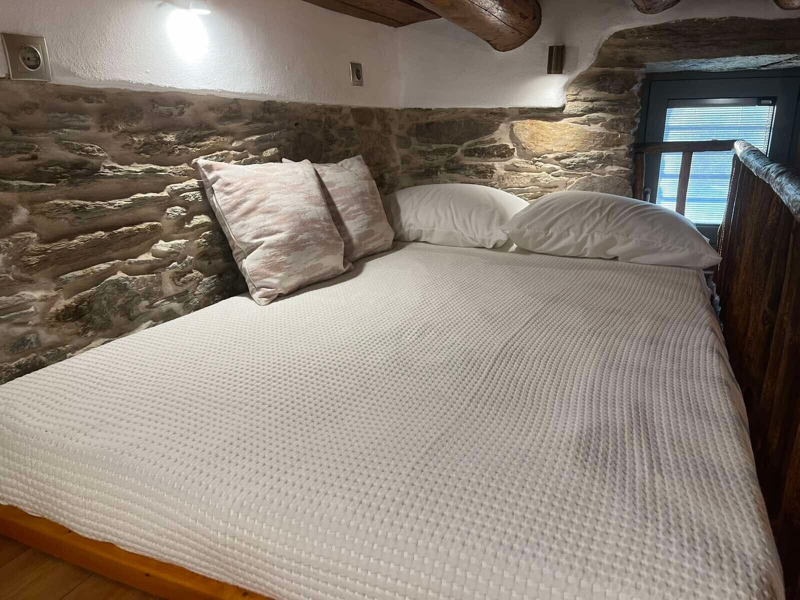 Una cama con una cubierta blanca texturizada, una pared de piedra y una viga de techo de madera.