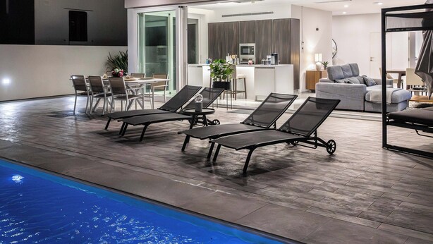 Una casa moderna con piscina, mobiliario exterior y una cocina visible a través de puertas de cristal.