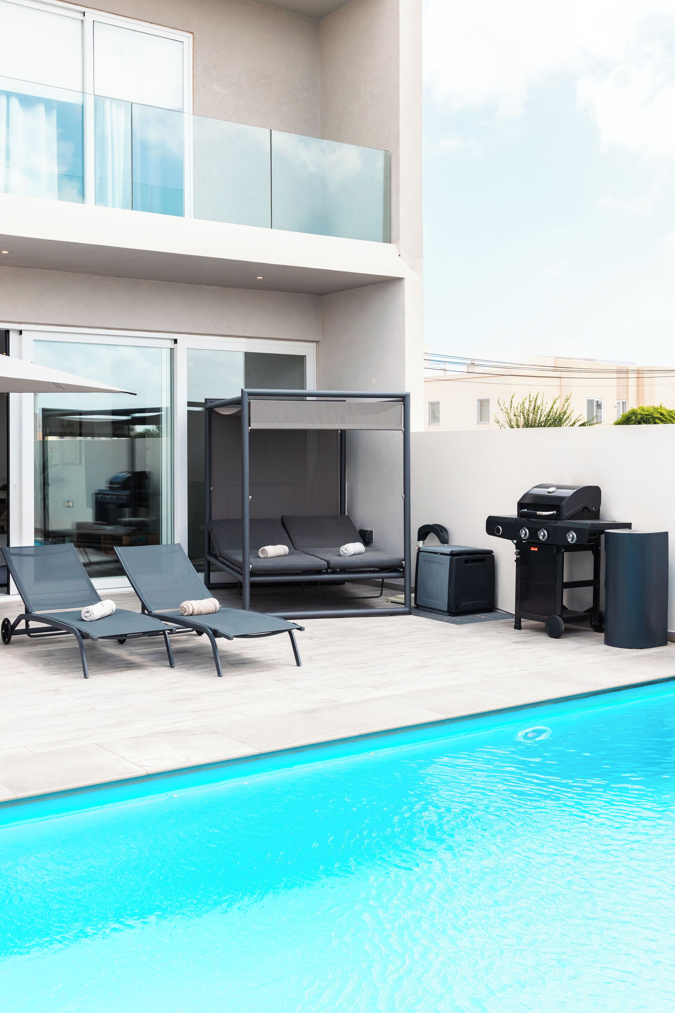 A modern outdoor pool area with lounge chairs, a grill, and a glass-enclosed seating area.