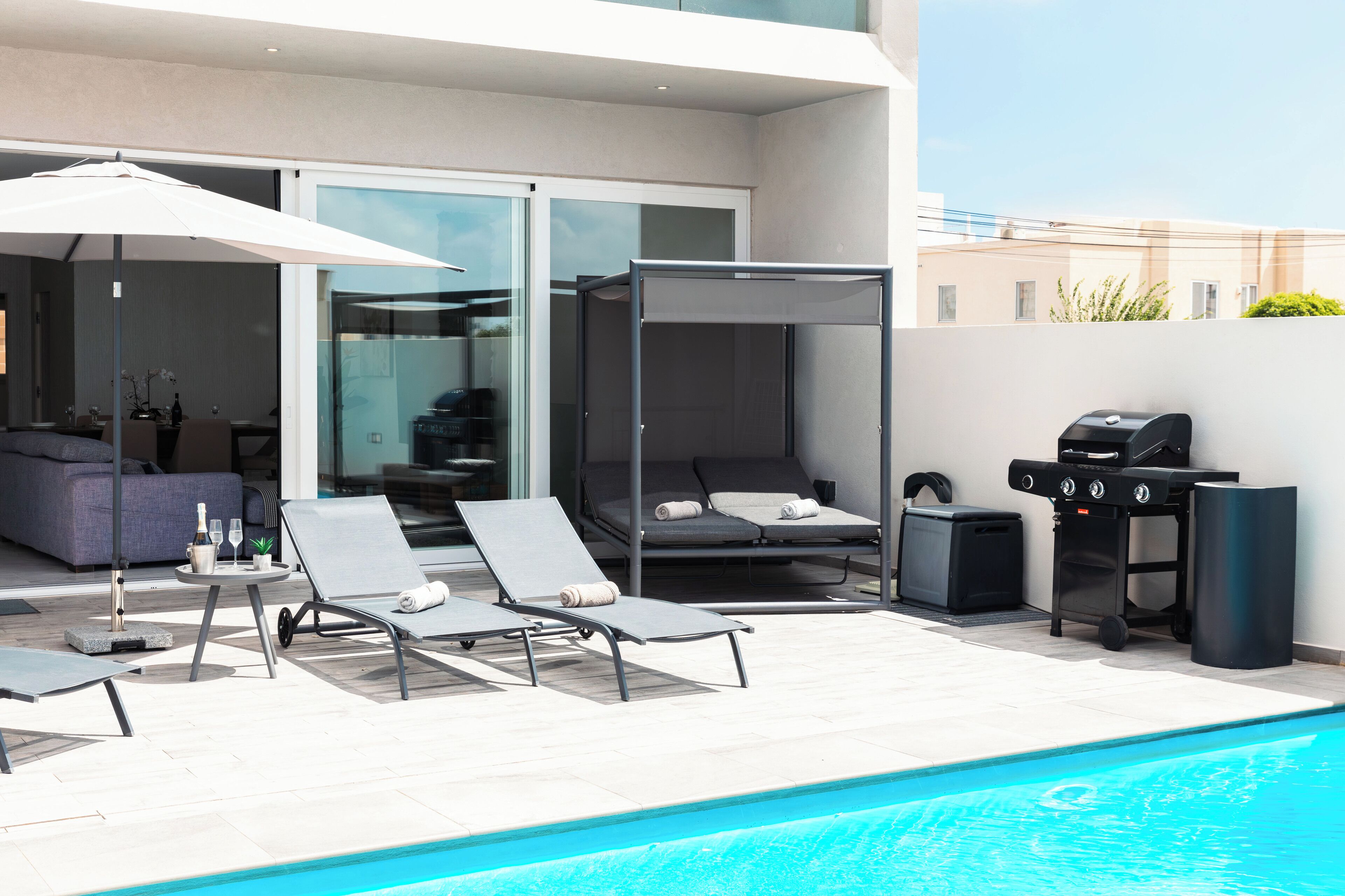 A modern outdoor pool area with lounge chairs, a grill, and a glass-enclosed patio.