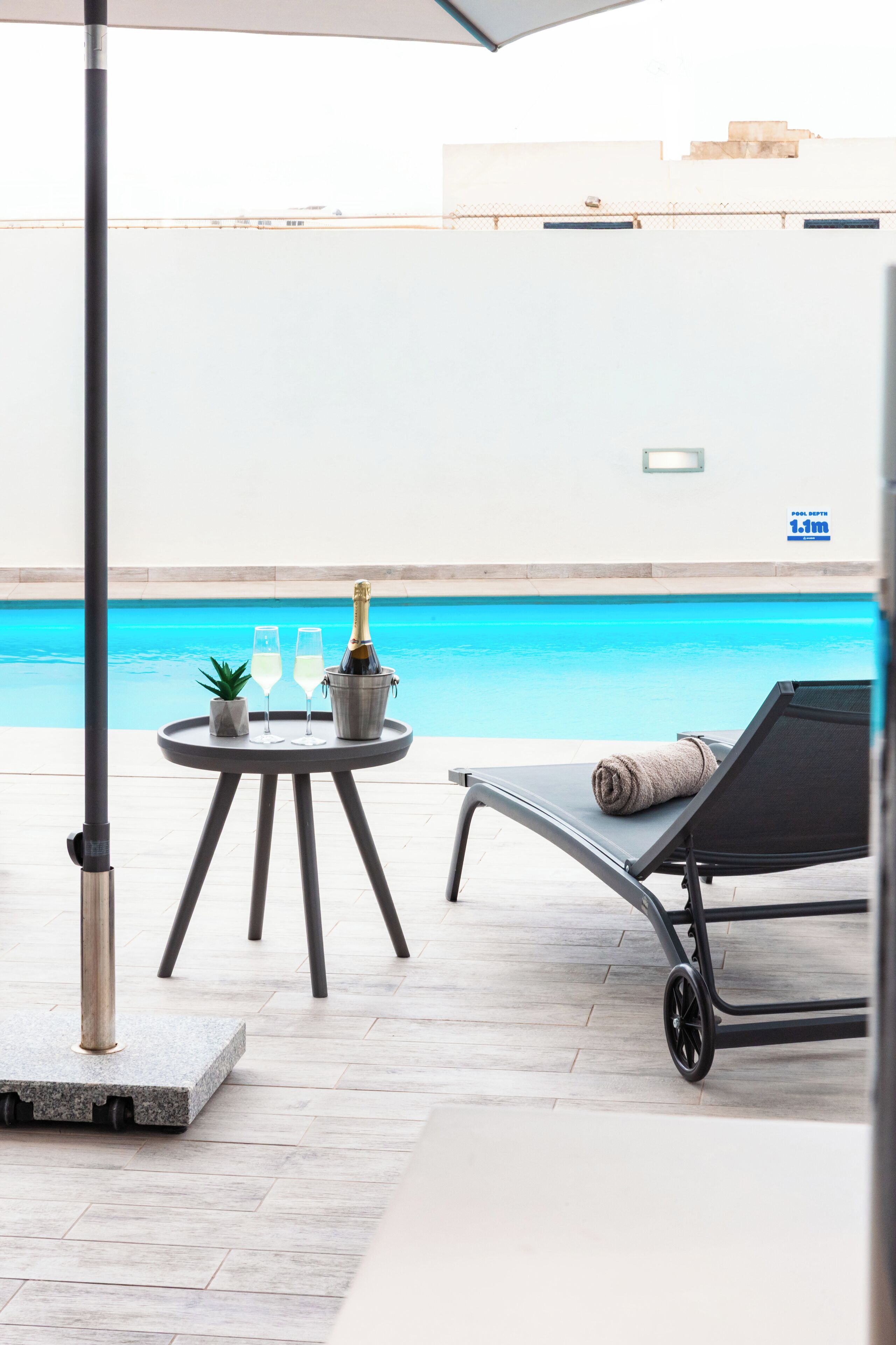 A poolside area with a table, two glasses, a bottle, a plant, and a lounge chair.