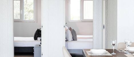 A modern hotel room with a bed, a small bedside table, a dining table with chairs, and a window.
