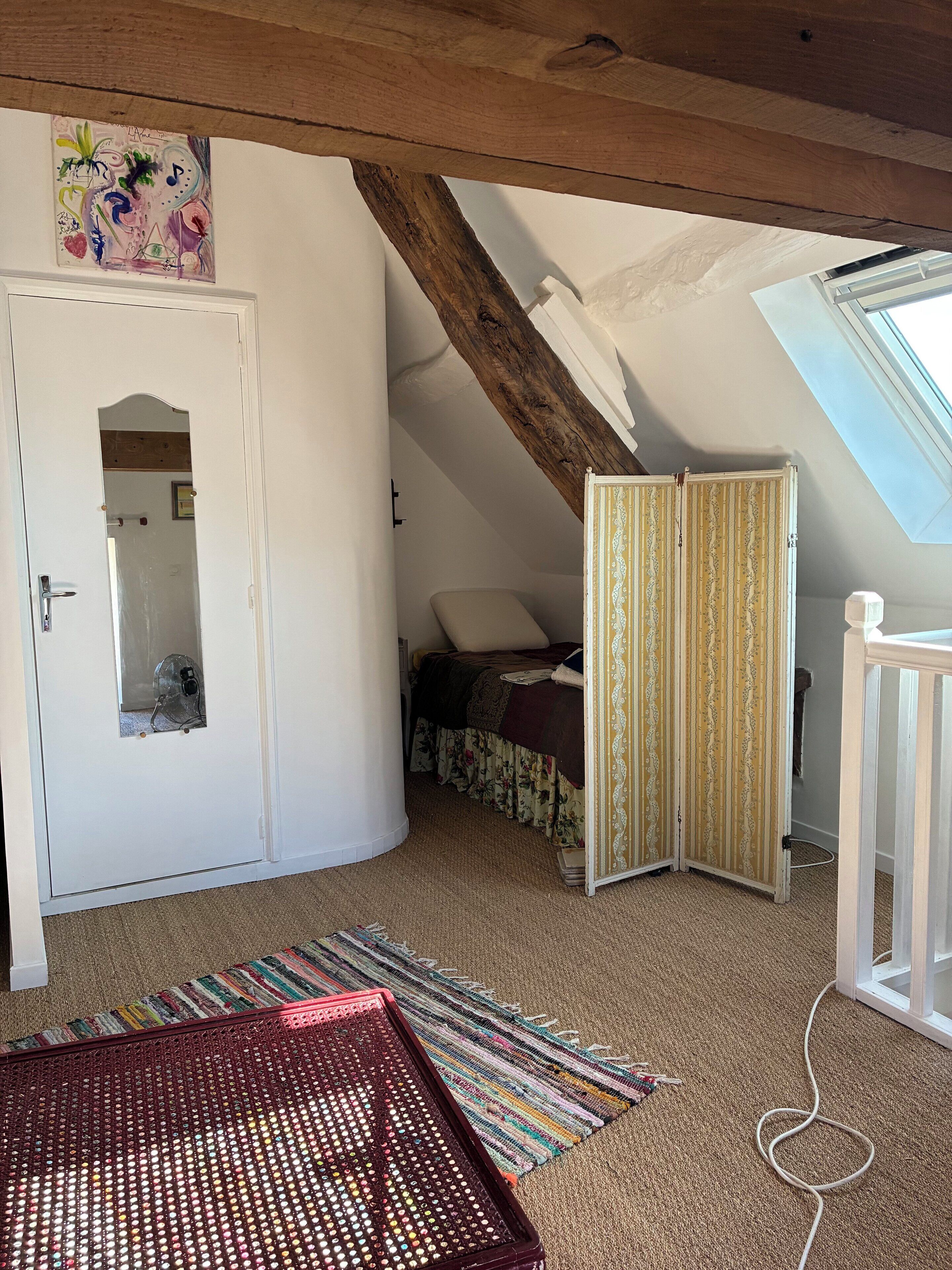 Ein Zimmer mit schräg steilendem Dach, einem Bett mit floralem Bettbezug, einem gemusterten Vorhang und einem kleinen Fenster.