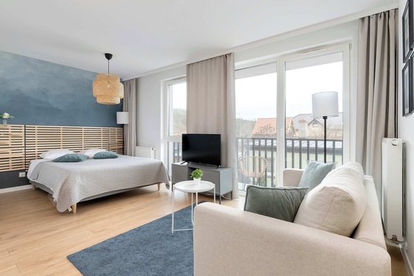 A bright studio-style living and sleeping area with a bed near large windows, a small sofa, a TV stand, and a balcony visible outside. Natural light fills the modern space with light wood flooring and neutral decor.
