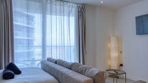 Een moderne hotelkamer met een groot bed, een bank, een staande lamp en uitzicht op de stad door dunne gordijnen.