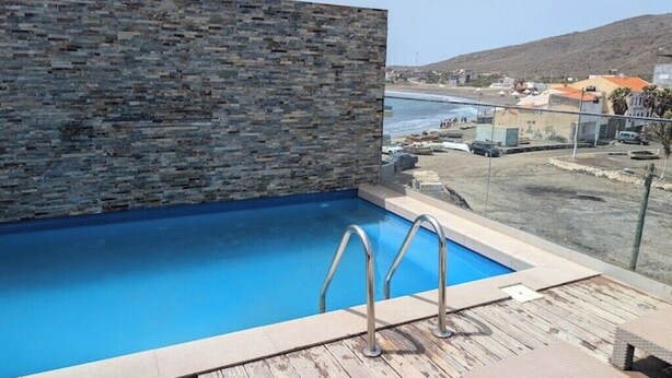 A swimming pool with a stone wall, a wooden deck, and a view of the sea and buildings. - Beachfront Bliss in Praia Baixo – 1Min Walk to Sea (Praia Baixo)