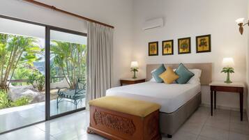A bedroom with a bed, two bedside tables, a bench, a sliding glass door to a balcony, and framed pictures on the wall.