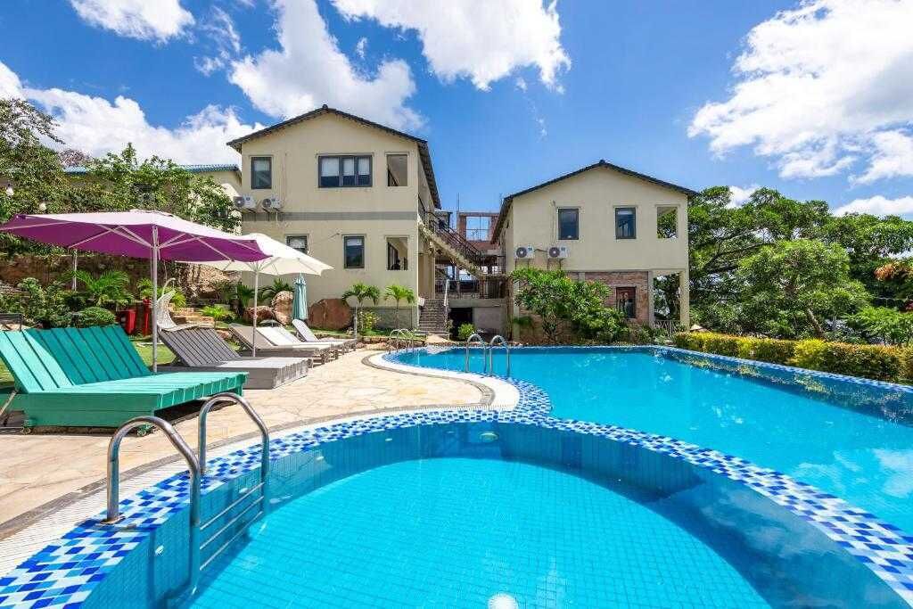 Une piscine dont le bord est recouvert de carrelage, entourée de chaises longues et de parasols, avec des bâtiments en arrière-plan.