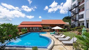 A swimming pool with lounge chairs and a covered seating area, surrounded by greenery and a building with a tiled roof.