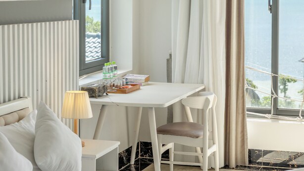 A hotel room with a bed, a desk, a chair, a lamp, and a view of the sea.