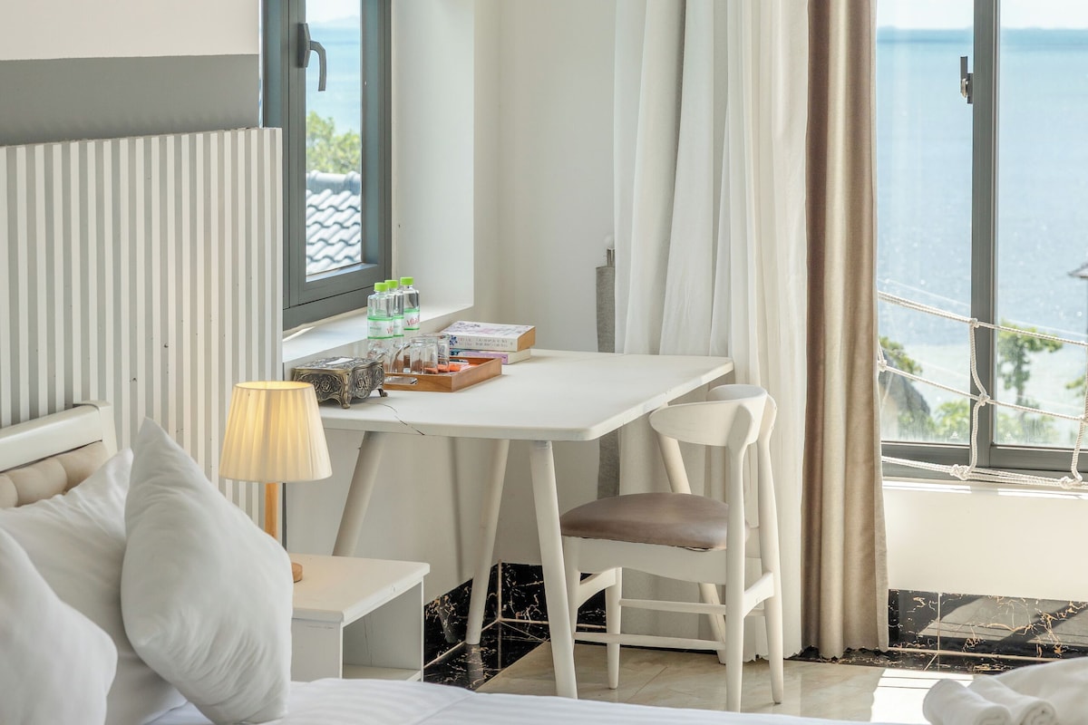 A hotel room with a bed, a desk, a chair, a lamp, and a view of the sea.