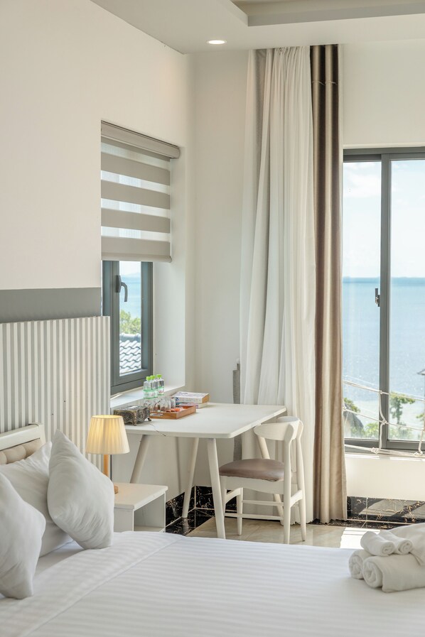 A hotel room with a bed, a desk, a chair, a lamp, and a view of the sea. - Palette Casa De Sol Beach Resort (Phu Quoc)