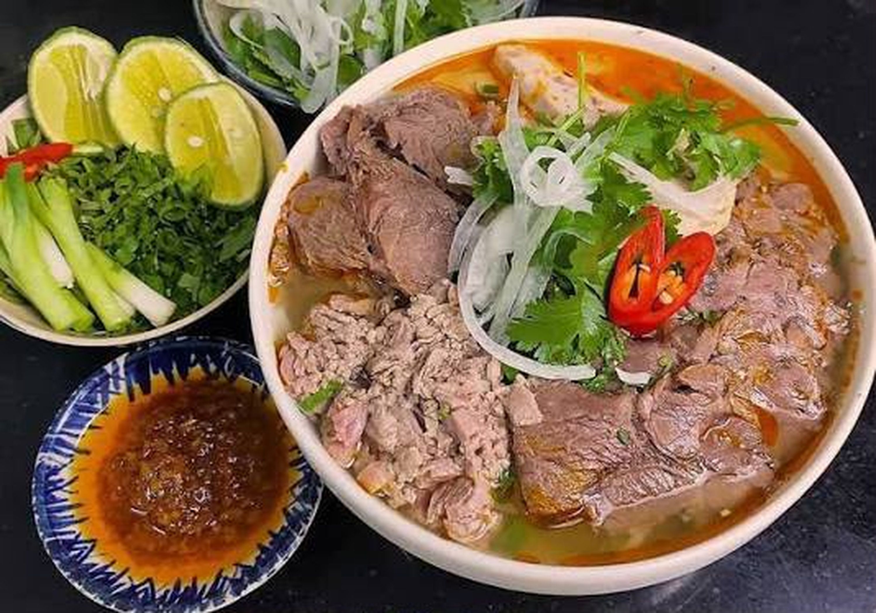 A bowl of soup with meat, garnished with herbs and vegetables, and a side of sliced cucumbers and a small bowl of sauce.