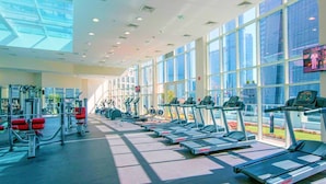 Et fitnesscenter med løbebånd, styrkemaskiner og et tv-skærm, der viser en gruppe mennesker.