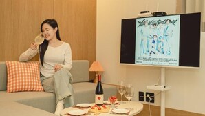 Una mujer sentada en un sofá con una botella de vino y copas sobre una mesa, un televisor mostrando a un grupo de personas, y una lámpara sobre la mesa.