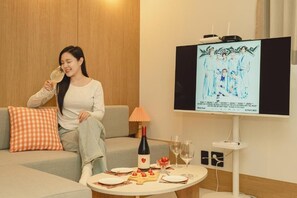 A woman sitting on a sofa with a wine bottle and glasses on a table, a TV displaying a group of people, and a lamp on the table. - HOTEL DAUN GURI (Guri)