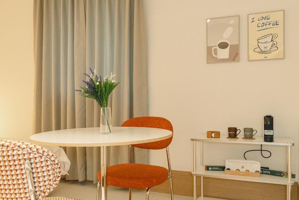 Une pièce avec une table ronde, deux chaises orange, un vase blanc contenant des plantes, une machine à café et deux tableaux encadrés accrochés au mur.