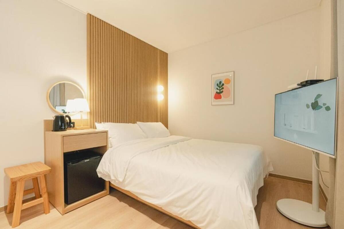 A hotel room with a bed, bedside table, TV, and a small stool.