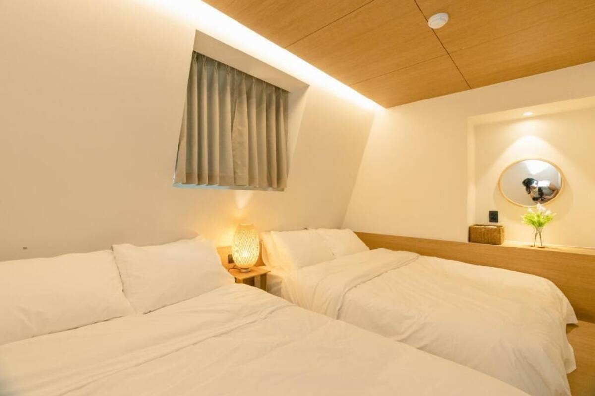 A hotel room with two beds, a window with curtains, a bedside table with a lamp, and a mirror on the wall.