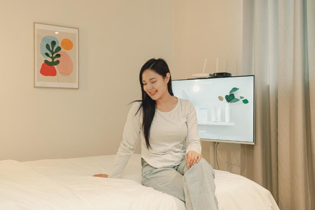 Eine Person sitzt auf einem Bett in einem Zimmer, in dem sich ein Fernseher an der Wand befindet und ein gerahmtes Bild darüber hängt.