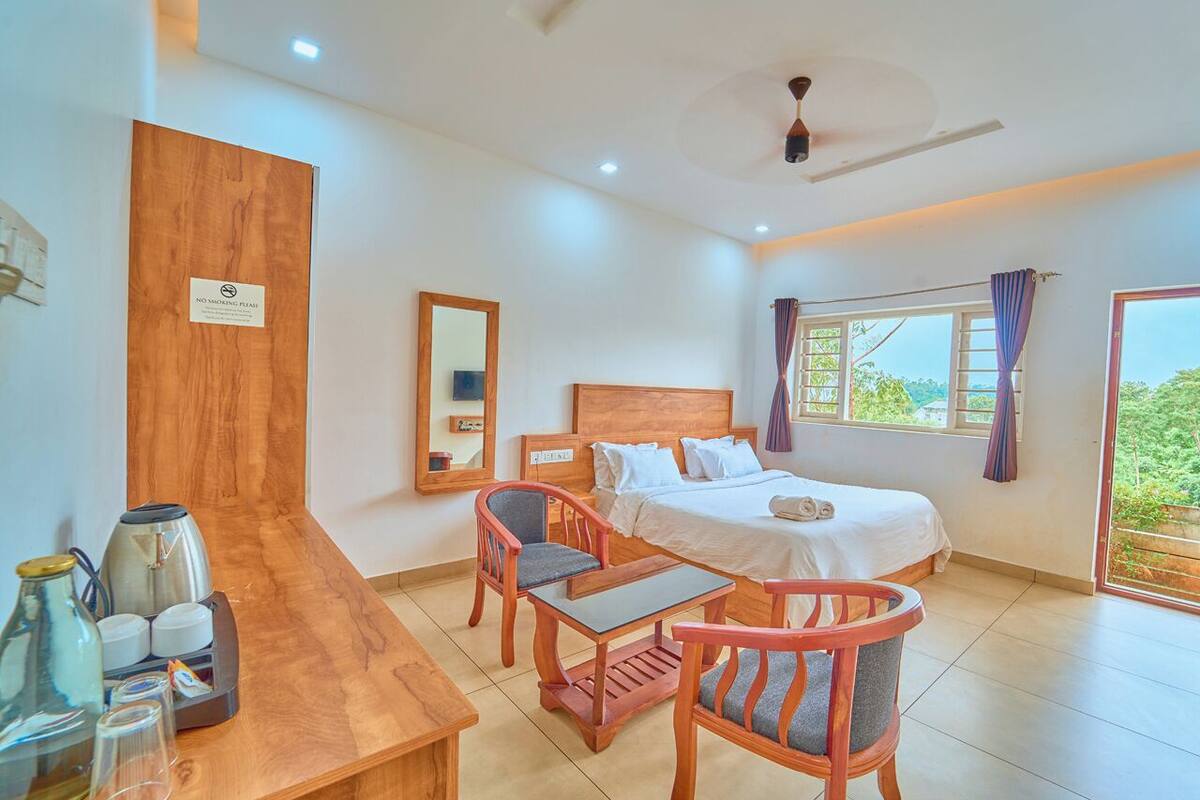A hotel room with a bed, a desk with a coffee maker, two chairs, a mirror, and a window with curtains.