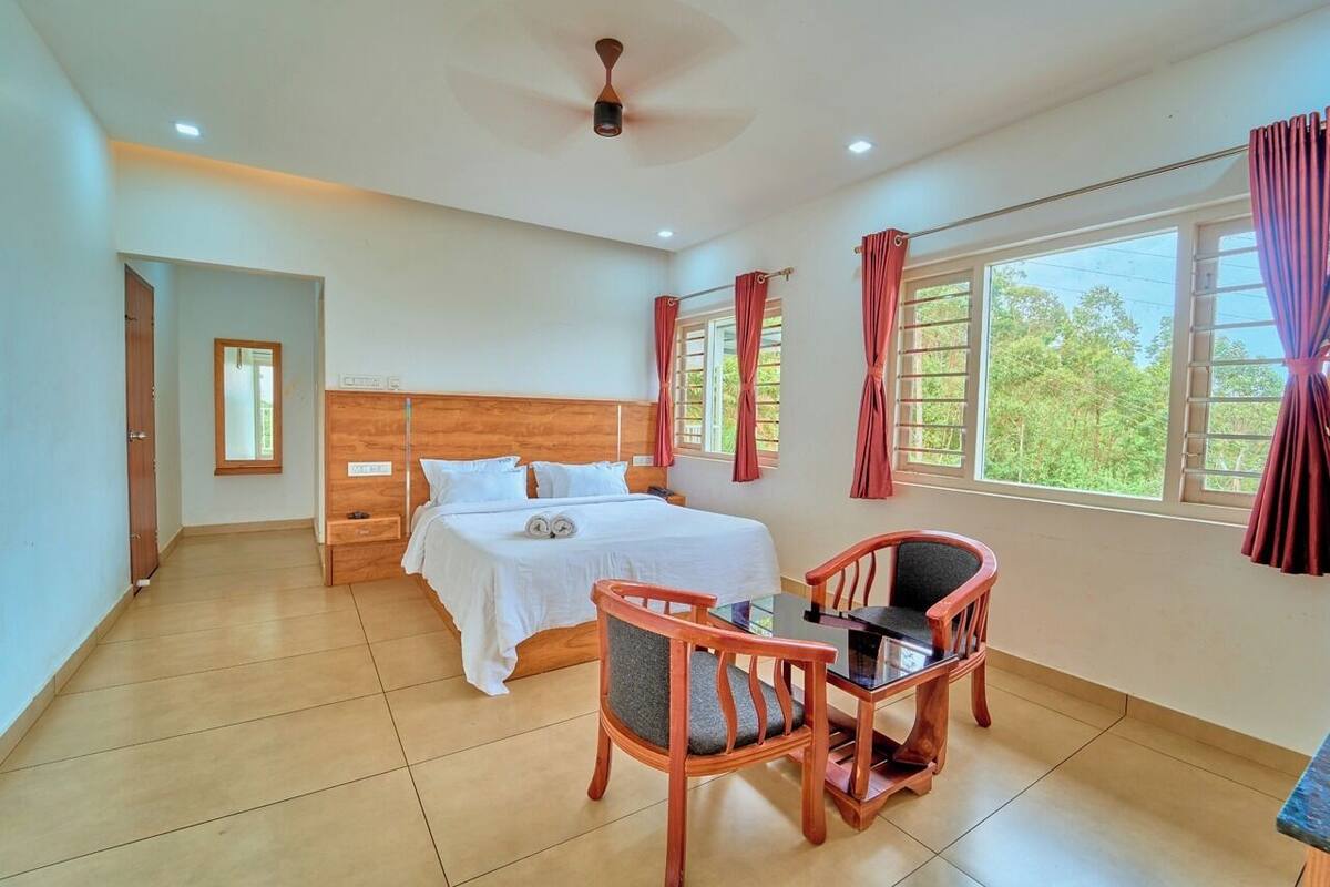 A bedroom with a bed, a desk, and chairs, a ceiling fan, and large windows with curtains.
