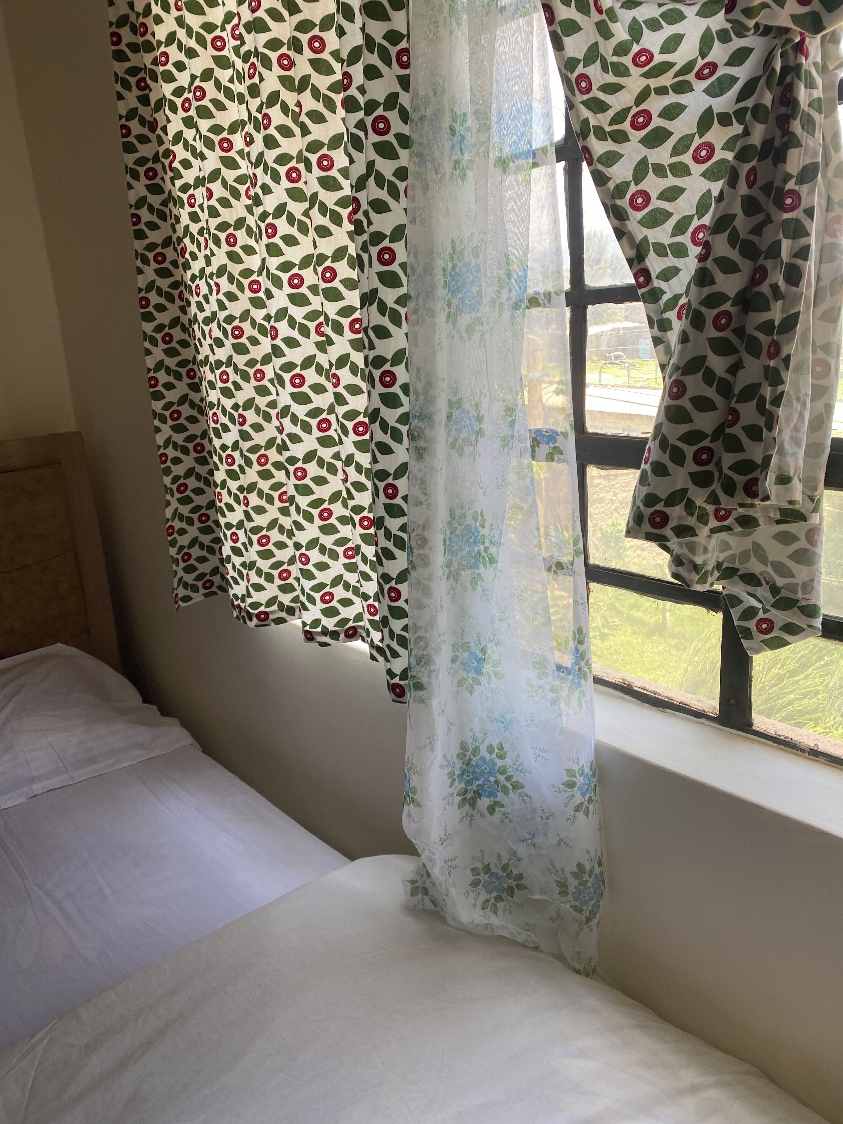Un lit avec un drap blanc, une fenêtre donnant sur un paysage verdoyant et des rideaux ornés d’un motif de feuilles.