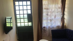 Una habitación con sofá, una mesita, una alfombra roja, televisor montado en la pared, una ventana con cortinas y una puerta.