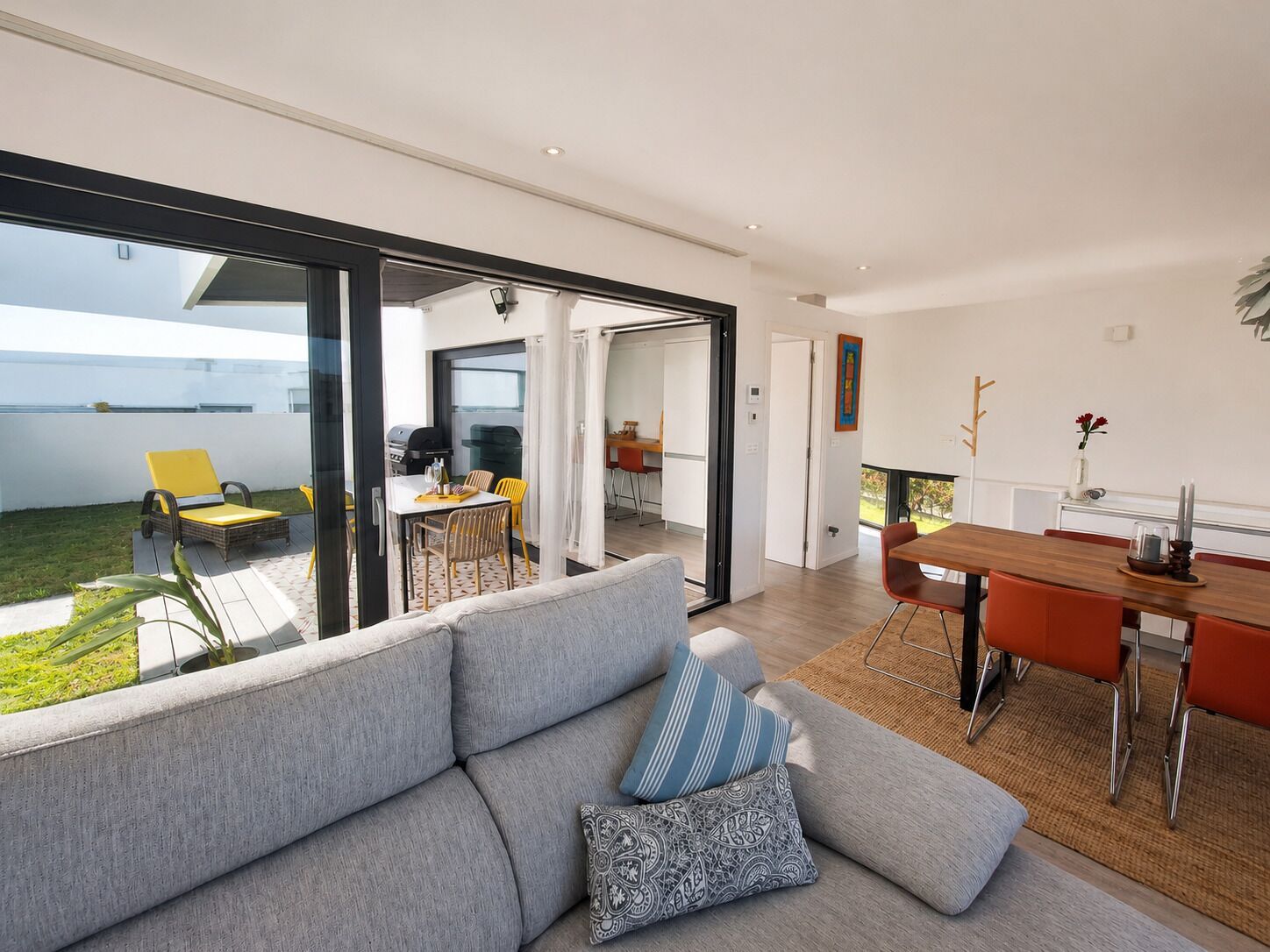 A modern living room with a grey sofa, a wooden dining table with orange chairs, and a glass sliding door leading to an outdoor area with a yellow chair and a barbecue grill.