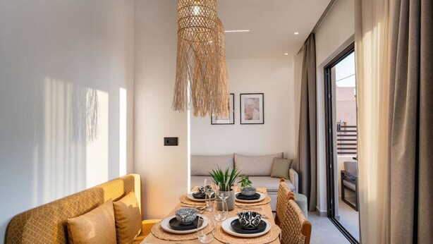 Un coin repas avec une table dressée pour deux personnes, un luminaire suspendu en fibres tressées et un canapé en arrière-plan.