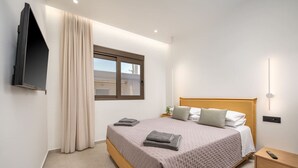 Une chambre à coucher comprenant un lit, un téléviseur fixé au mur, une fenêtre avec des rideaux, une table de chevet et une commode.