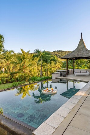 Piscina cristalina con un pabellón y zona de estar exterior, rodeada de exuberante vegetación y palmeras.