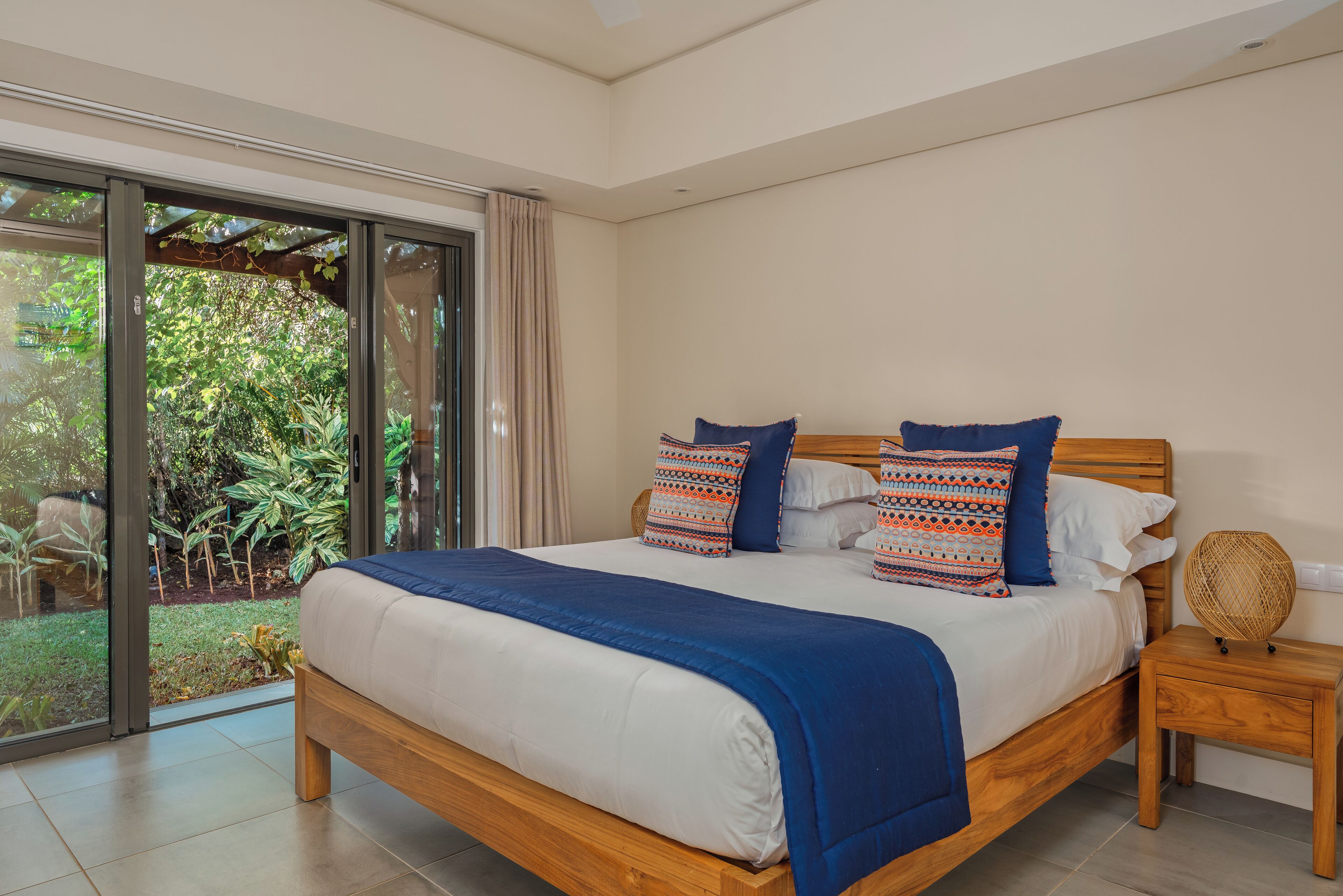 Una camera da letto con un letto grande, una testiera in legno, un comodino e vista su un giardino attraverso una porta scorrevole in vetro.