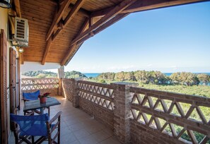 Un’area esterna riparata con tavolo e sedie, un muro in mattoni con decorazioni a griglia, e vista sul mare e sulla vegetazione.