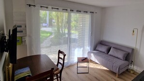 Een kamer met een houten eettafel, een grijze bank en een raam met dunne gordijnen.