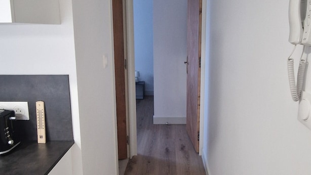 Een smalle gang met houten vloeren, witte muren en een deur naar een andere kamer.