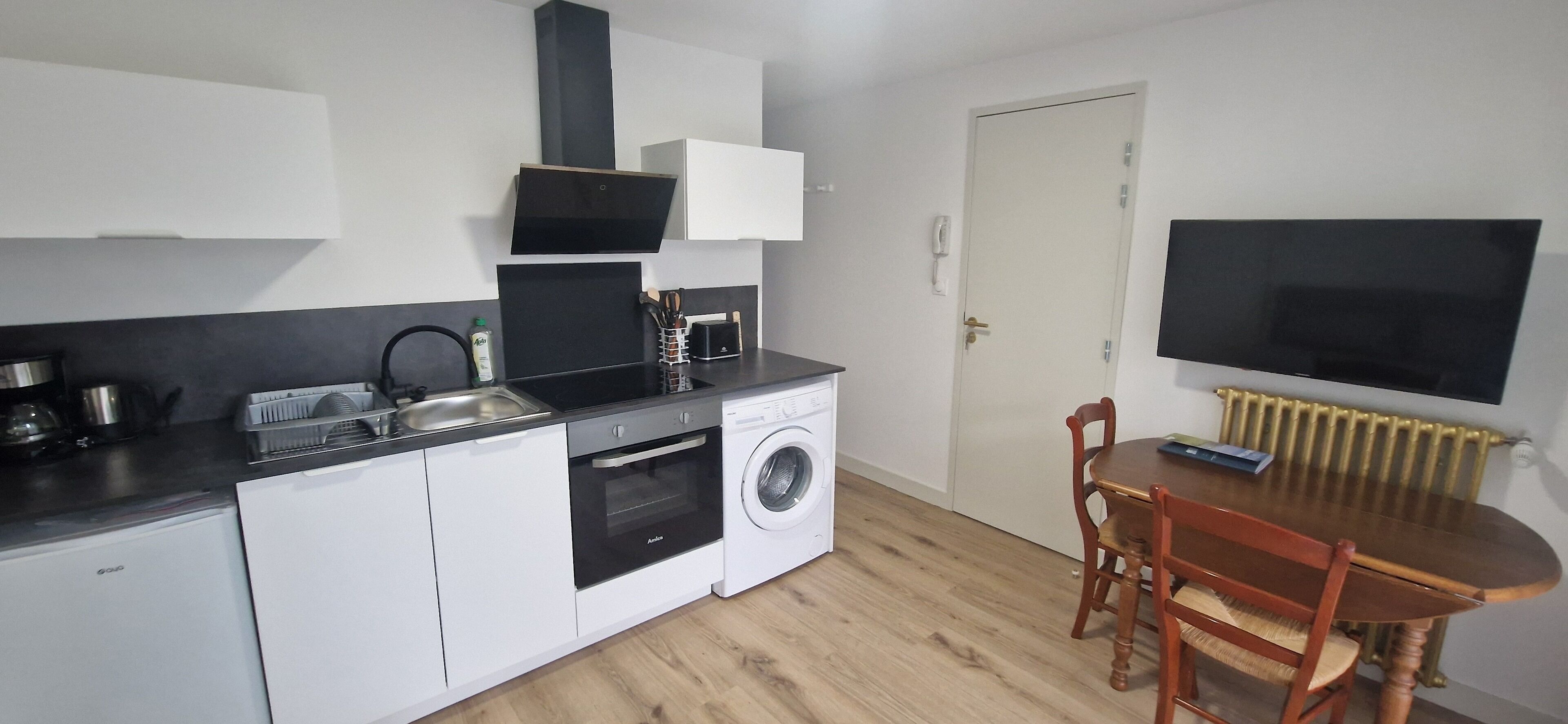 A compact kitchen with a washing machine, a dining table with chairs, and a flat-screen TV mounted above the table.