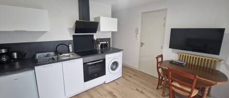 A compact kitchen with a washing machine, a dining table with chairs, and a flat-screen TV mounted above the table.