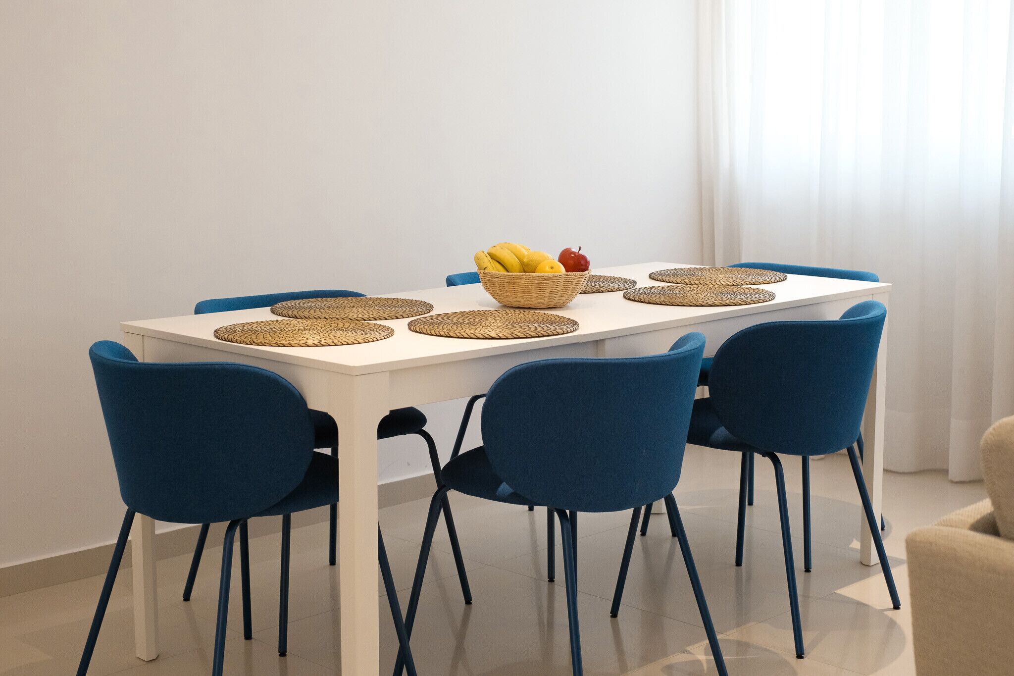 A dining table with a woven placemat, a bowl of fruit, and blue chairs.