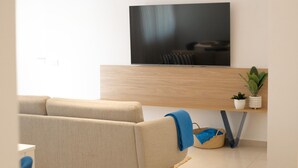 A modern living room with a flat-screen TV mounted on a wooden wall, a beige sofa, a small table with a plant, and a basket. - Nila 1 by Huskalia - City Center (Los Llanos de Aridane)