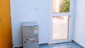 A room with a door, a small refrigerator, and a window with a view of a paved area and greenery.