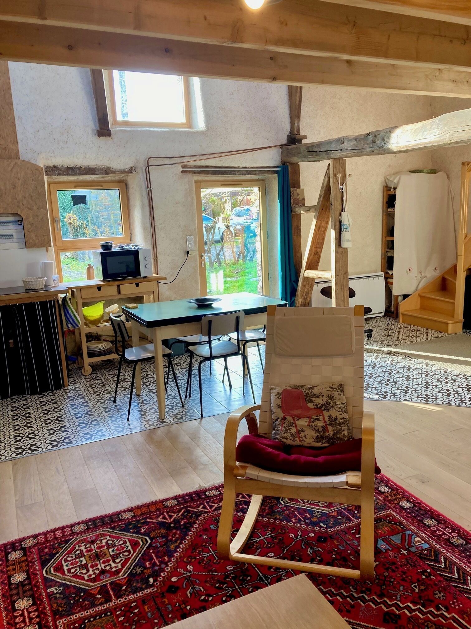 Een kamer met een eettafel, stoelen, een rood tapijt met patroon, een houten stoel met kussen en een raam met uitzicht.